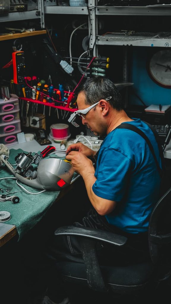 Technician with magnifying glasses working on electronic repairs in a cluttered workshop.